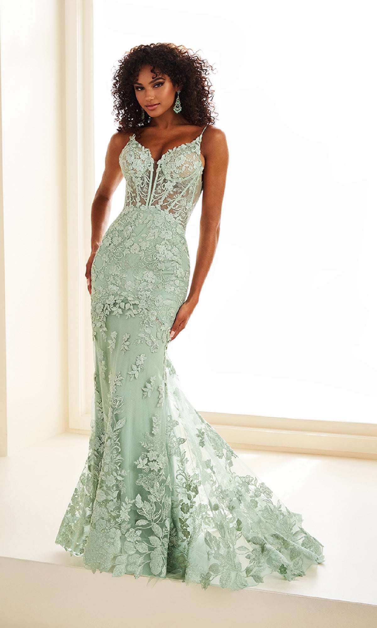 A woman stands in front of a bright window wearing the Ellie Wilde EW36014, a fitted, floor-length mint green lace mermaid prom dress with a sheer corset bodice and floral appliqué.